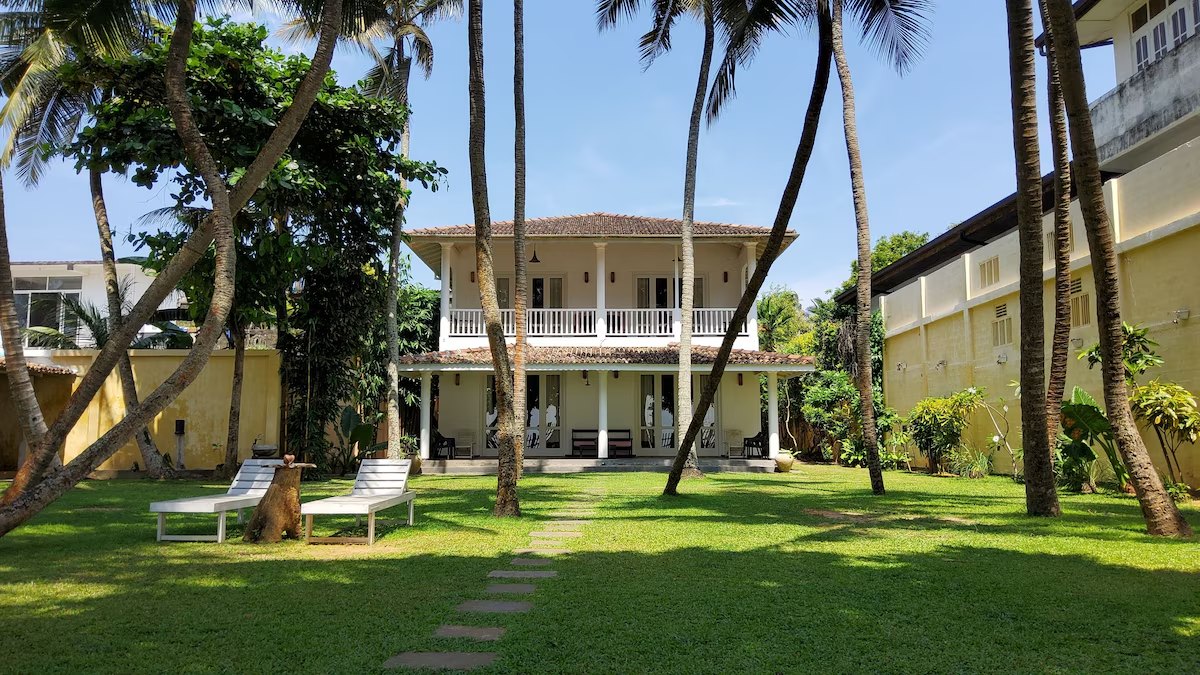 Villa Inlet Galle — the renovated two-storey villa seen from the garden, with the lawn, path and coconut palms
