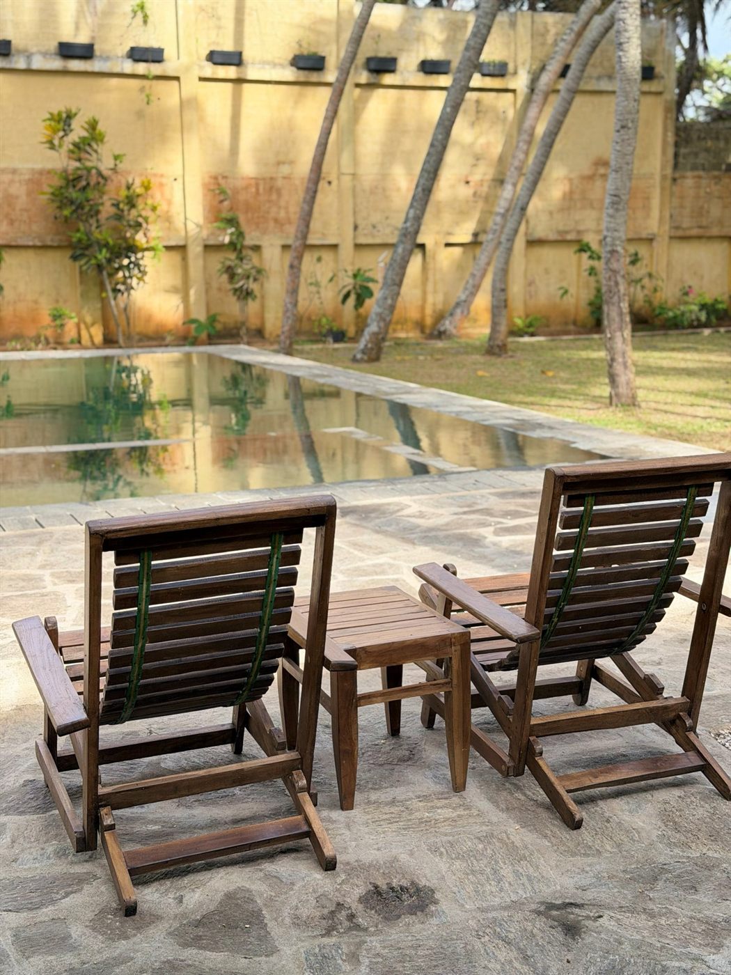 Two teak chairs and a small table set out beside the pool in the afternoon light